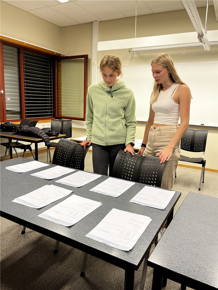 To jenter bak en pult dekket av oppgaver - Klikk for stort bilde