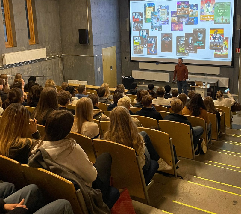Et auditorium, fullt av elever, avbildet skrått bakfra med foredragsholder Sigbjørn Mostue foran ved lerretet.