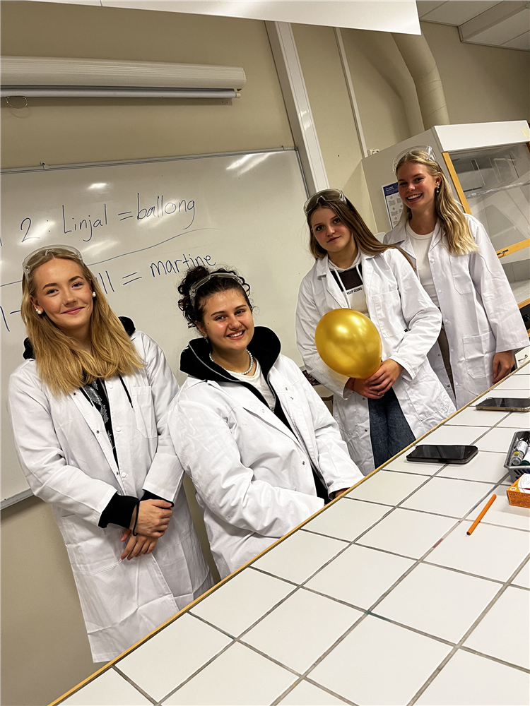 Fire jenter ikledd labfrakker forran tavla - Klikk for stort bilde